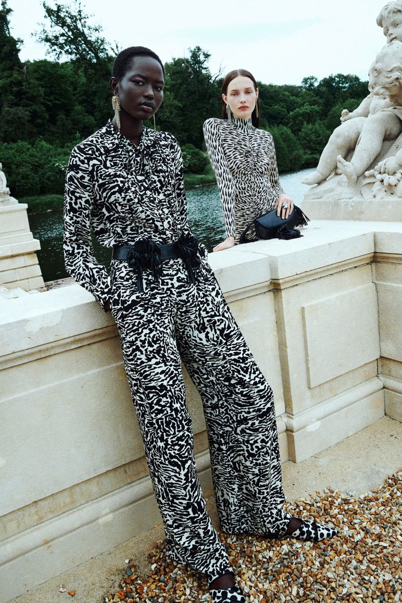 Two fashion models pose on a stone ledge wearing black-and-white print outfits with a river and trees in the background.