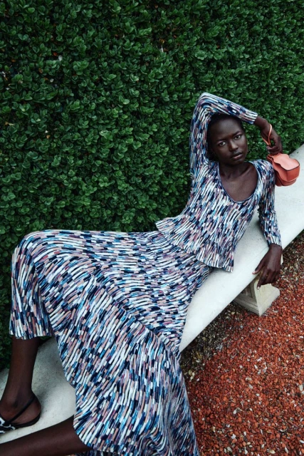 An African woman model poses reclining outdoors in a patterned dress with a purse, against a green hedge and a stone bench.