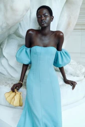 Model in off-the-shoulder blue dress holding a yellow handbag against a sculpted white stone background