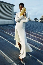 Model standing on a metal rooftop wearing a white long-sleeve dress and holding a yellow clutch bag
