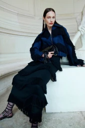 Model Clara Denison seated wearing a navy and black fringe jacket with a matching long tiered fringe skirt and strappy sandals.