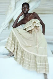 Model Nyaduola Gabriel poses in a tiered beige ruffled maxi dress while seated in front of a white classical marble sculpture.
