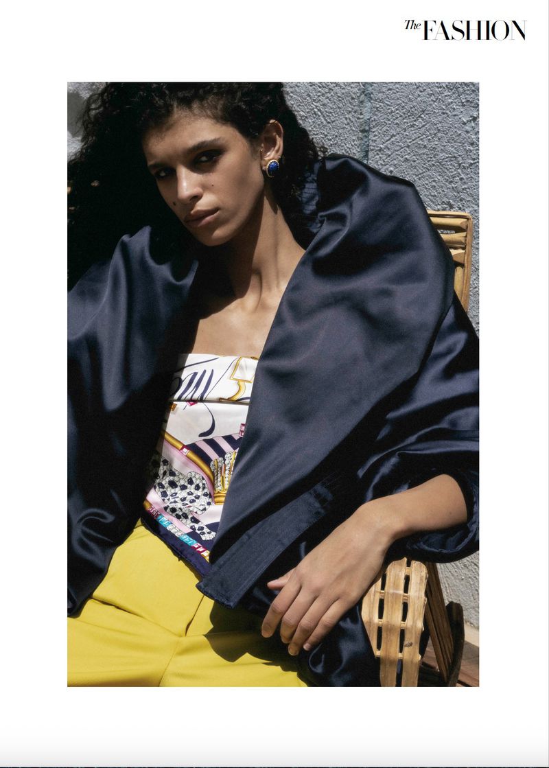 Model seated outdoors in a wicker chair wearing a voluminous dark navy satin wrap, graphic printed top, and bright yellow trousers.
