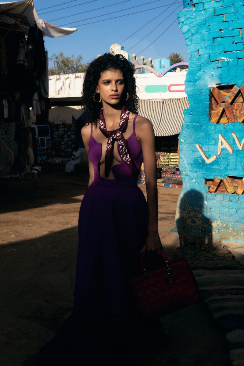 Model wearing a purple gown with a neck scarf and holding a handbag beside a textured blue painted brick pillar outdoors.