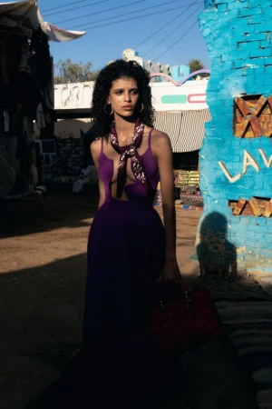 Model wearing a purple gown with a neck scarf and holding a handbag beside a textured blue painted brick pillar outdoors.