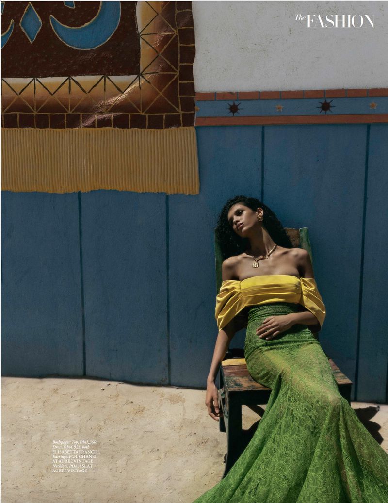 Model reclining in a wooden chair wearing a yellow off-the-shoulder top and vibrant green lace maxi skirt against a blue painted wall.