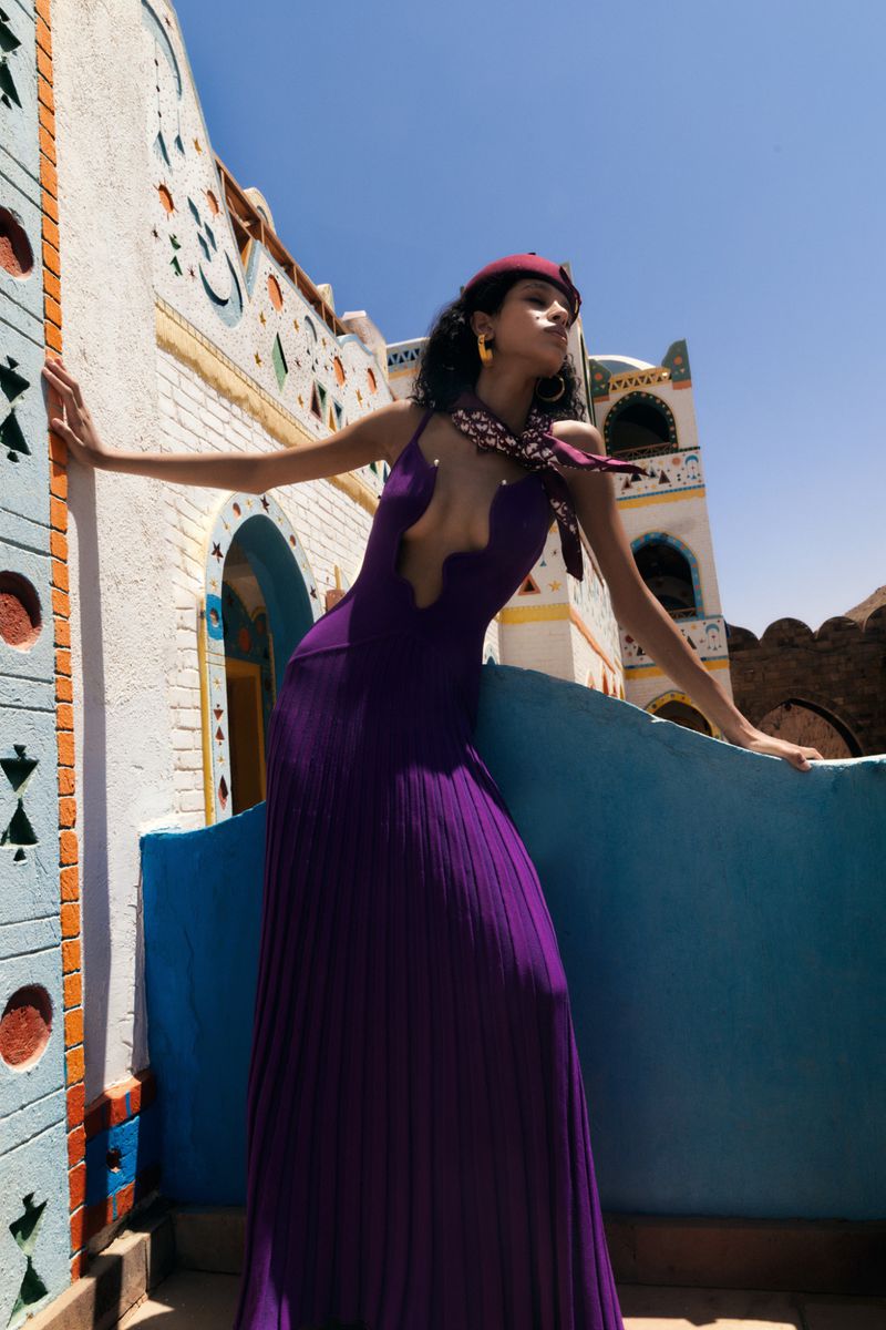 Model in a long purple pleated gown and maroon beret leaning against a blue wall in front of painted architecture under a clear sky.