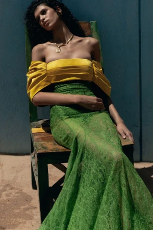 Fashion model in a yellow off-the-shoulder top and long green lace skirt seated on a wooden chair against a blue wall in direct sunlight.