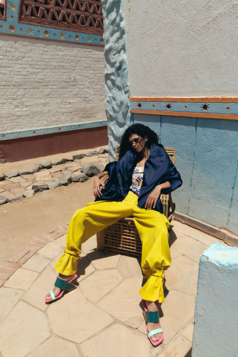 Woman in bright yellow wide-leg trousers, navy blue shawl, and sunglasses reclining in a wicker chair on a sunlit stone patio.