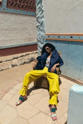Woman in bright yellow wide-leg trousers, navy blue shawl, and sunglasses reclining in a wicker chair on a sunlit stone patio.