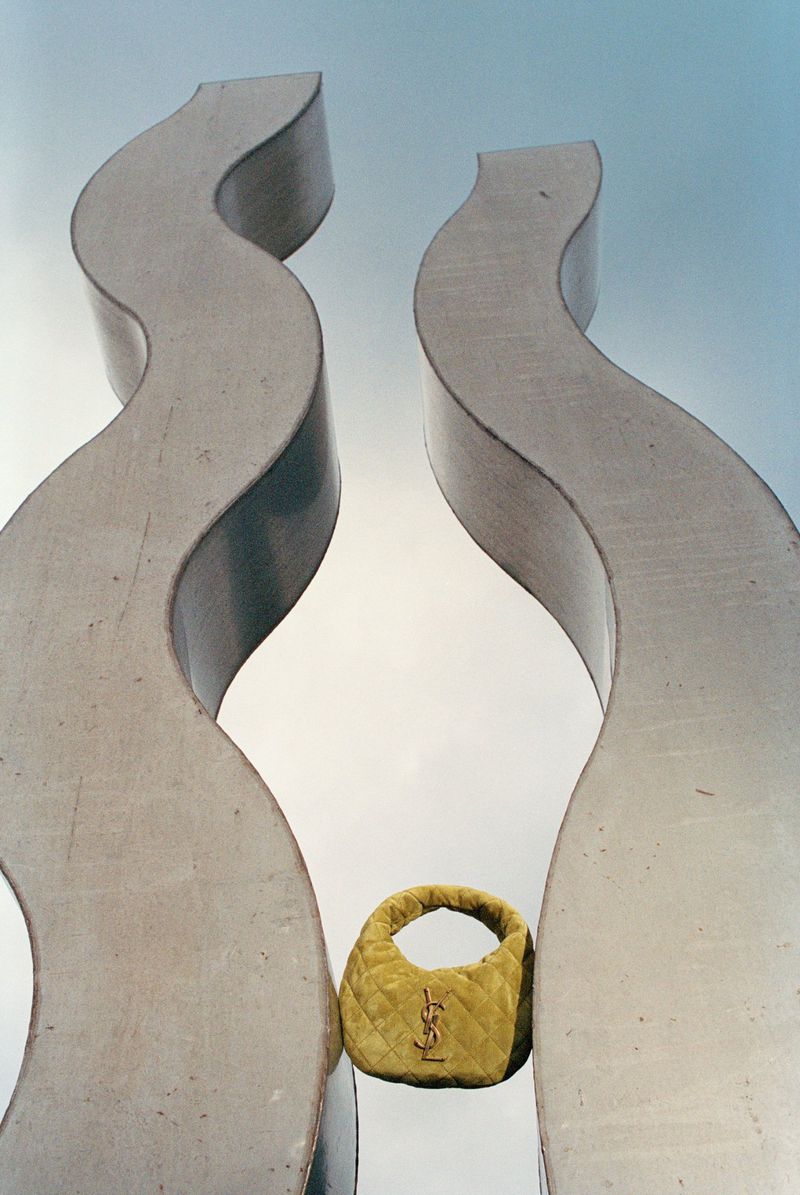Quilted yellow mini handbag wedged between two tall, curving sculptural concrete pillars under a pale sky.