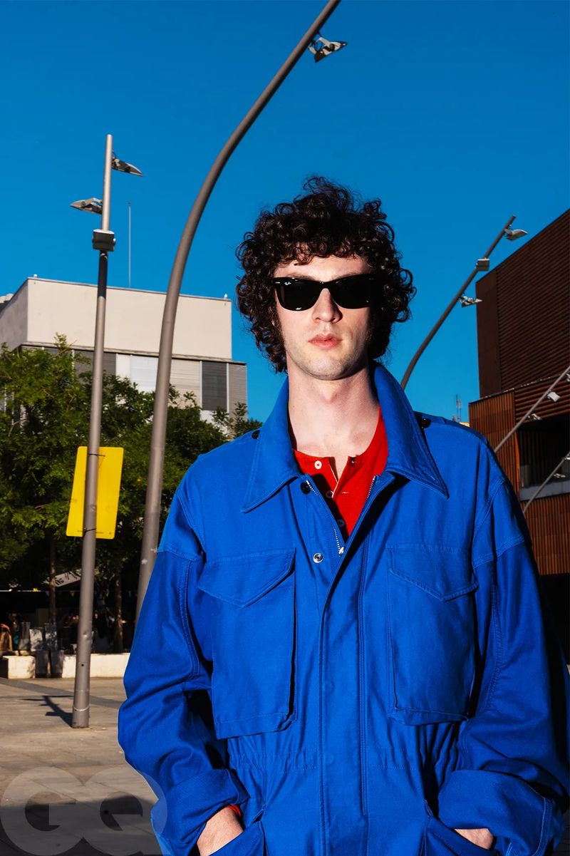 Dominic Sessa wearing dark sunglasses and a vibrant royal blue jacket outdoors under a bright blue sky.