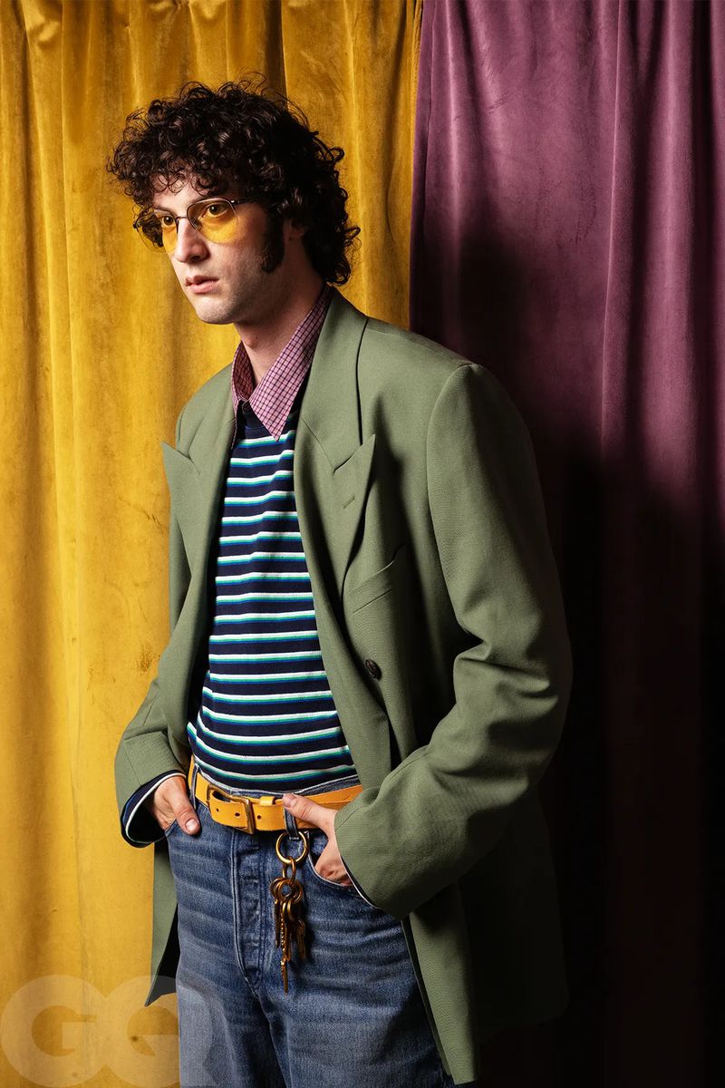 Actor Dominic Sessa wearing an olive green blazer, striped shirt, and yellow sunglasses in front of draped curtains.