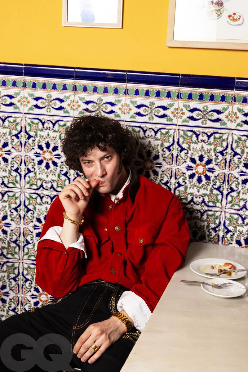 Dominic Sessa seated at a table wearing a red overshirt and gold bracelets against a patterned blue and white tiled wall.