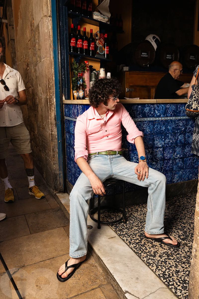 Dominic Sessa seated on a stool outside a bar wearing a pink polo shirt, light-wash jeans, and black flip-flops.