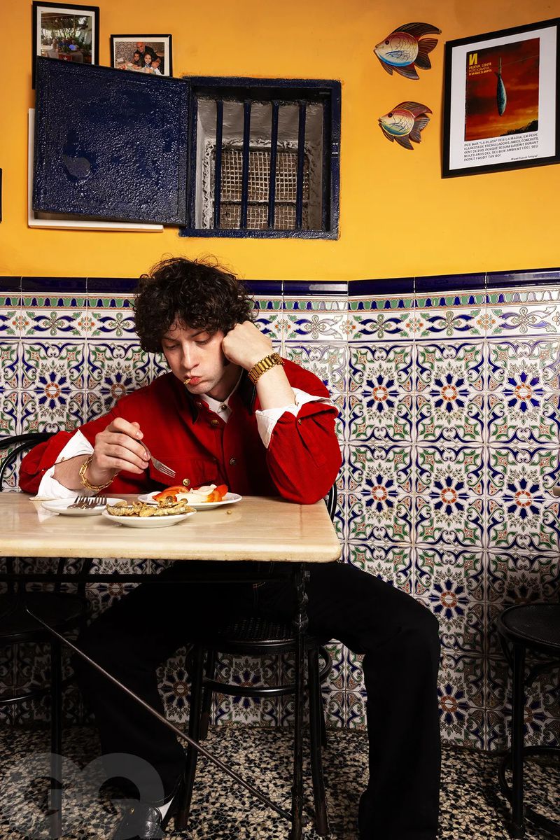 Dominic Sessa seated at a table eating in a red shirt and black trousers against a patterned tiled and yellow wall.