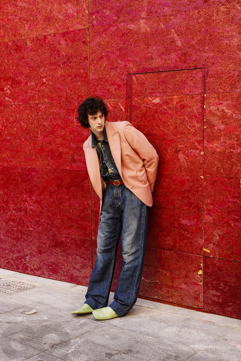 Actor Dominic Sessa posing in a peach blazer, wide-leg blue jeans, and light green shoes against a textured red wall.