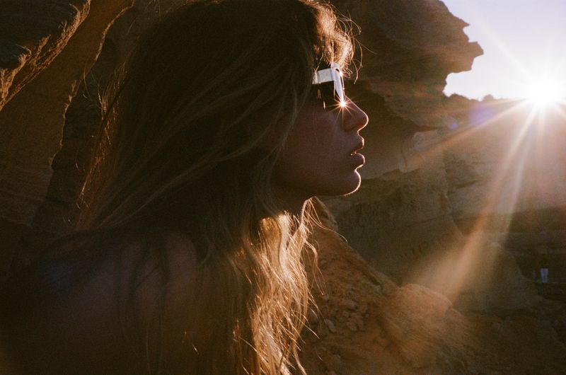 Woman with long hair and sunglasses poses near rock formations at sunset with lens flare creating a warm and sunny atmosphere.