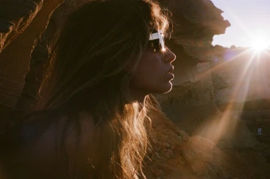 Close-up profile of a woman wearing futuristic sunglasses against a sunlit desert rock formation with intense lens flare.