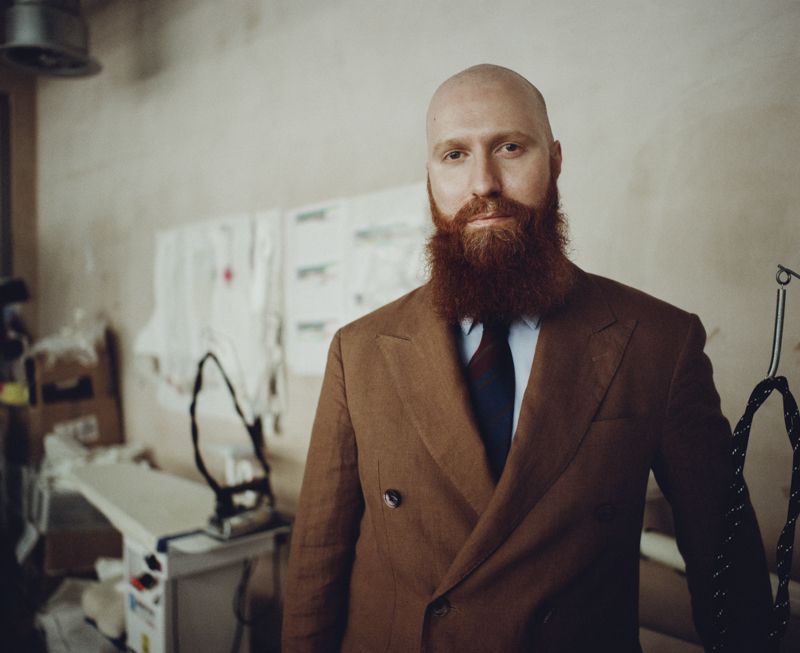Portrait of designer Michael Stewart with a full red beard wearing a brown double-breasted suit in a fashion studio.