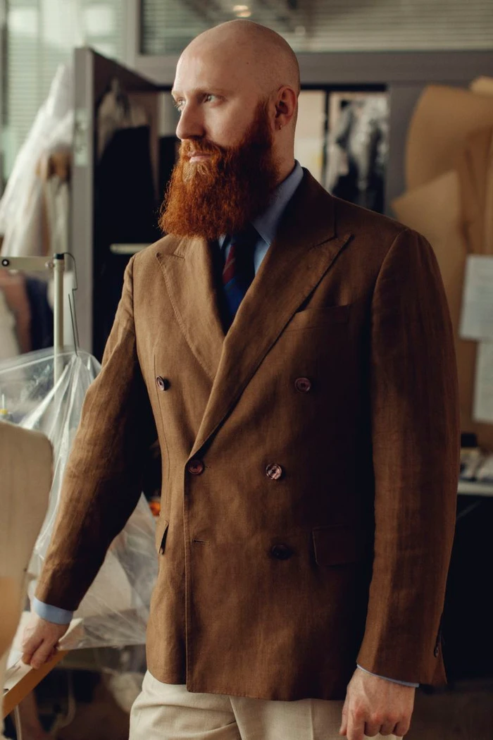 Bald man with a prominent red beard wearing a brown double-breasted tailored blazer in a fashion design studio.