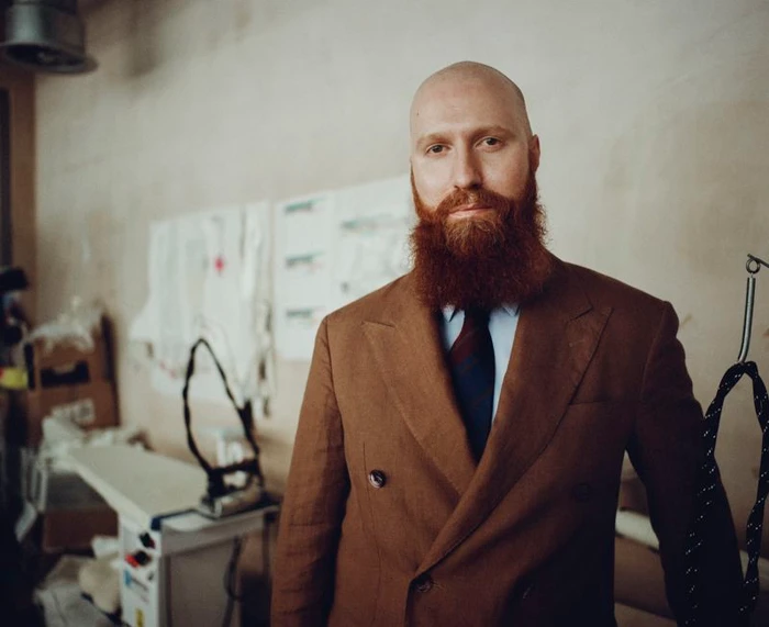 Portrait of designer Michael Stewart with a full red beard wearing a brown double-breasted suit in a fashion studio.