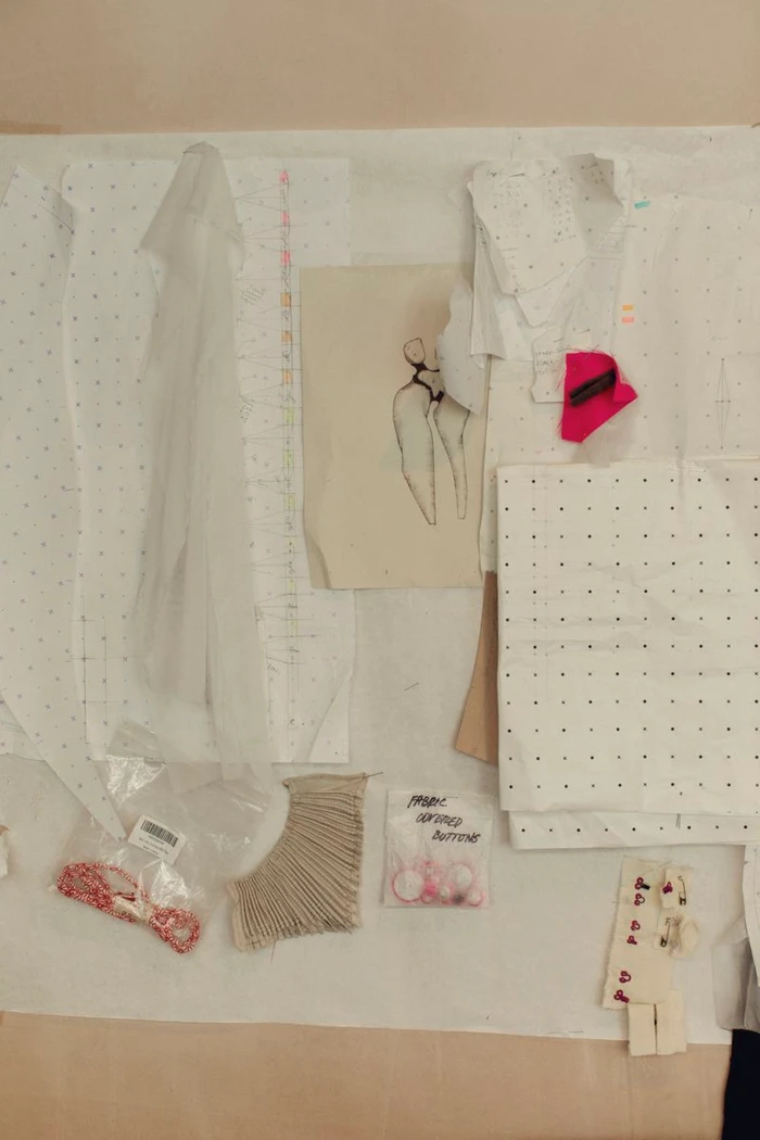 Overhead flat lay of fashion pattern paper, a figurative ink sketch, sheer fabric, pleated trim, and notions on a design work surface.