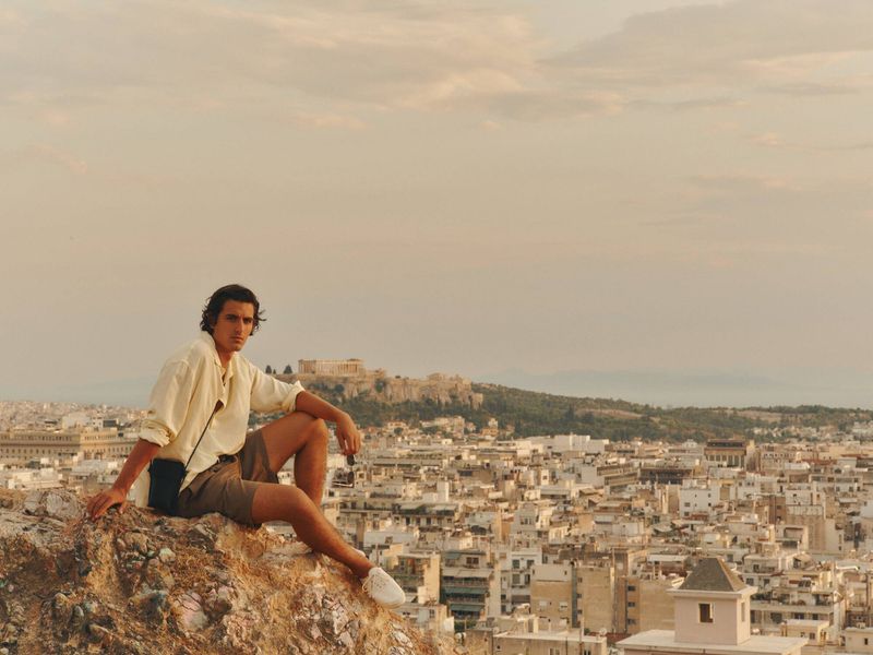 Man in a cream button-down shirt and brown shorts seated on a rocky hill overlooking an expansive cityscape with ancient ruins.