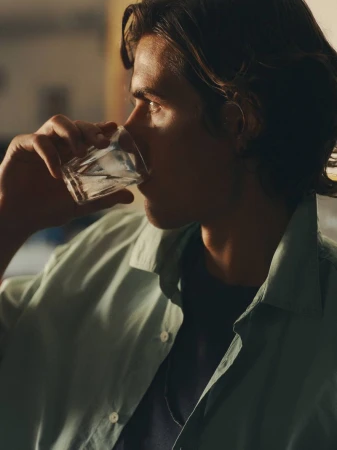 Man in profile drinking from a clear glass, wearing an unbuttoned sage green shirt over a dark t-shirt.