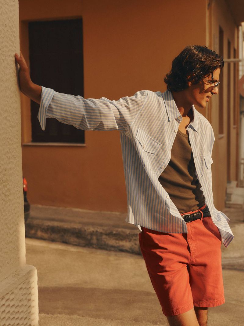 Model in a striped button-down shirt and coral shorts leaning against a textured wall on a sunlit street.