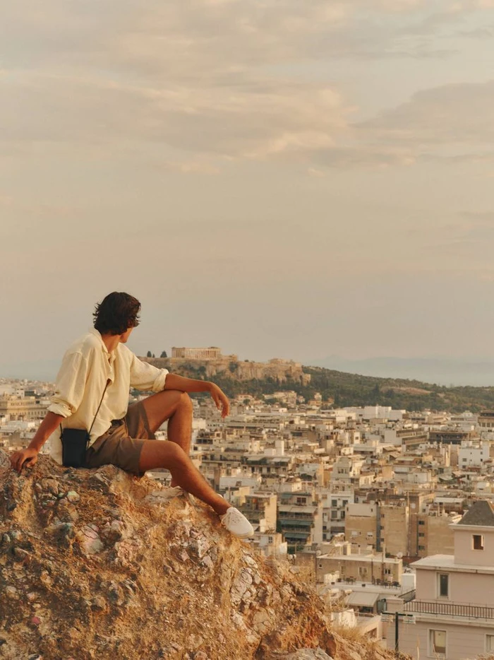 Man in a cream button-down shirt and brown shorts sitting on a rocky outcrop overlooking a sprawling sunny city with ancient ruins.