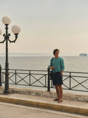 A man in a striped teal long-sleeve top and navy shorts stands on a seaside promenade beside an ornate lamppost overlooking the water.