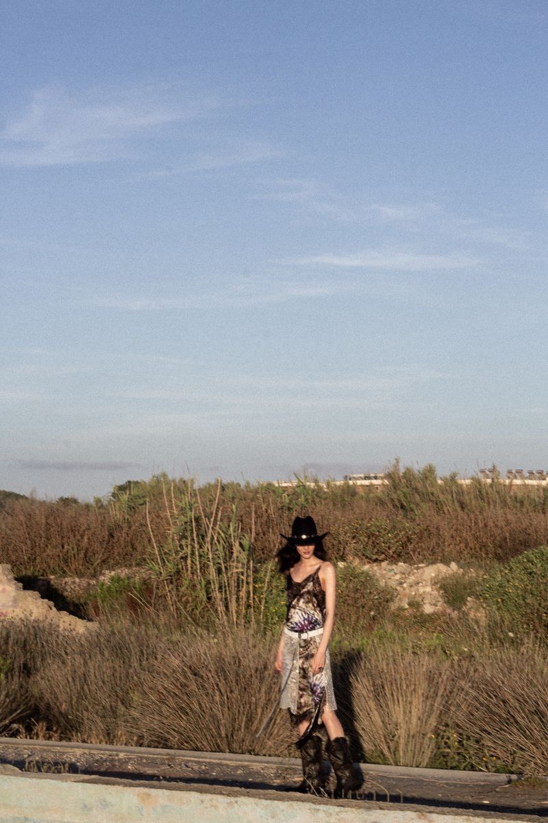 Model Vanessa Pompucci in a black cowboy hat and patterned dress standing in a dry grassy field under a pale blue sky.