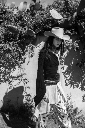 Model Vanessa Pompucci wearing a wide-brim cowboy hat, black long-sleeve top, and patterned trousers outdoors in bright sunlight.