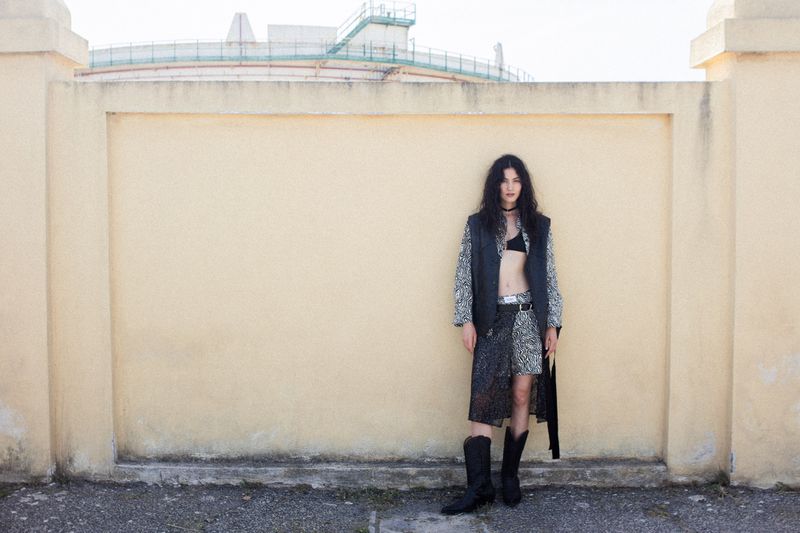 Model Vanessa Pompucci in a black leather vest, patterned ensemble, and cowboy boots standing against a pale wall.