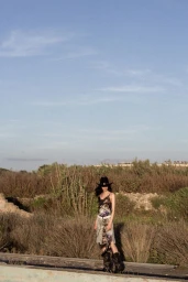 Model Vanessa Pompucci in a black cowboy hat and patterned dress standing in a dry grassy field under a pale blue sky.