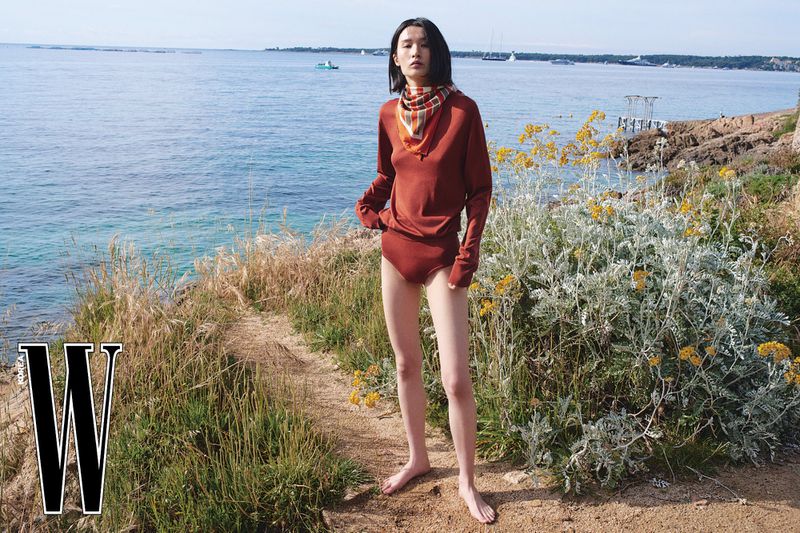 Model Esther Kim poses barefoot on a coastal path overlooking the blue sea in a rust knit top and patterned scarf for W Korea.