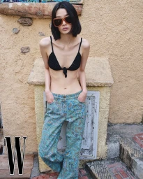Model wearing a black bikini top and patterned blue trousers leaning against a textured stucco wall outdoors.