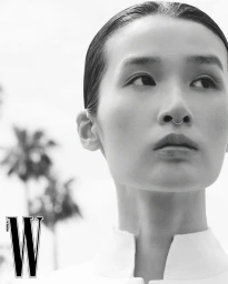 Black and white close-up portrait of model Esther Kim wearing a septum ring and white high-collar garment against blurred palm trees.