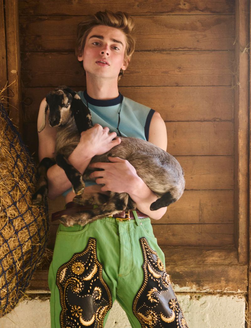 Model Luka Hoogedeure holds a small goat wearing a blue sleeveless top and green embroidered trousers against a wooden barn wall.