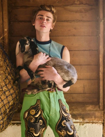 Model Luka Hoogedeure holds a small goat wearing a blue sleeveless top and green embroidered trousers against a wooden barn wall.