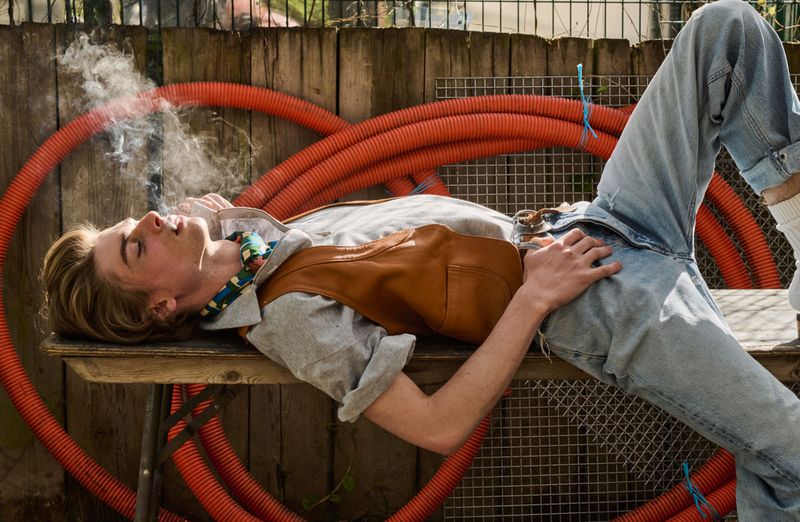 Model Luka Hoogedeure reclining on a wooden bench while smoking, wearing a brown leather vest and light wash denim jeans.