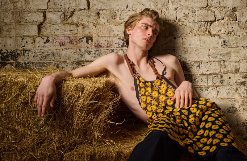 Model Luka Hoogedeure wearing a yellow patterned apron piece while reclining against hay bales and an exposed brick wall.
