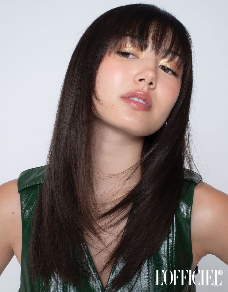 Portrait of actress Miku Martineau with straight brown hair and bangs wearing a shiny green vest against a white studio background.