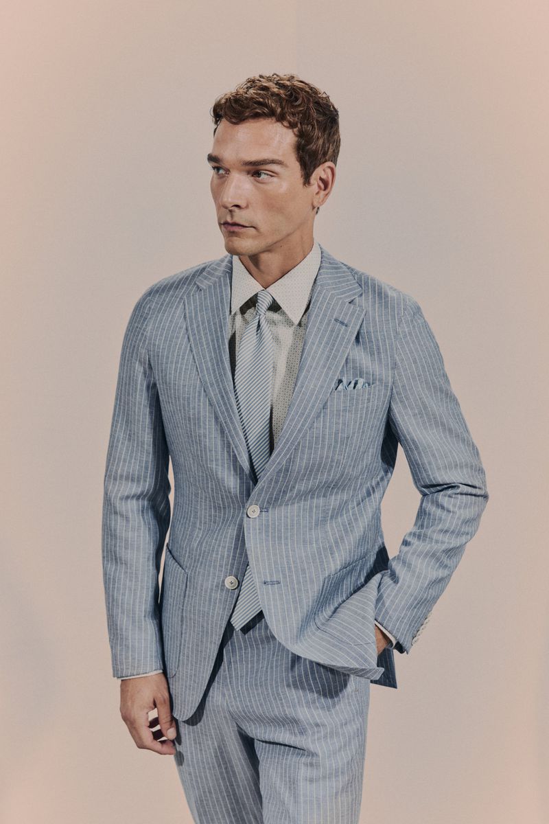A handsome adult man wearing a blue and white striped suit and tie poses against a light backdrop in a portrait fashion shot.