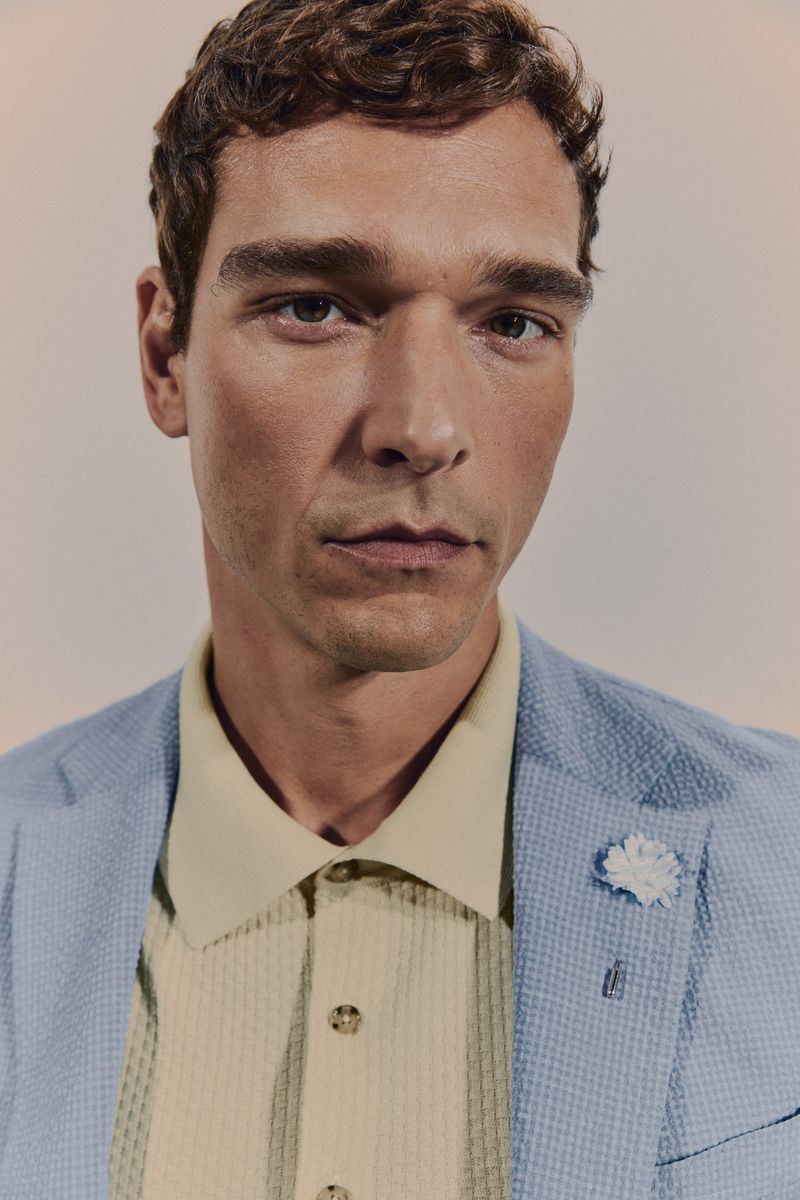 Close-up of a man wearing a light blue suit jacket over a collared shirt with a flower lapel pin. Portrait shot in neutral lighting.