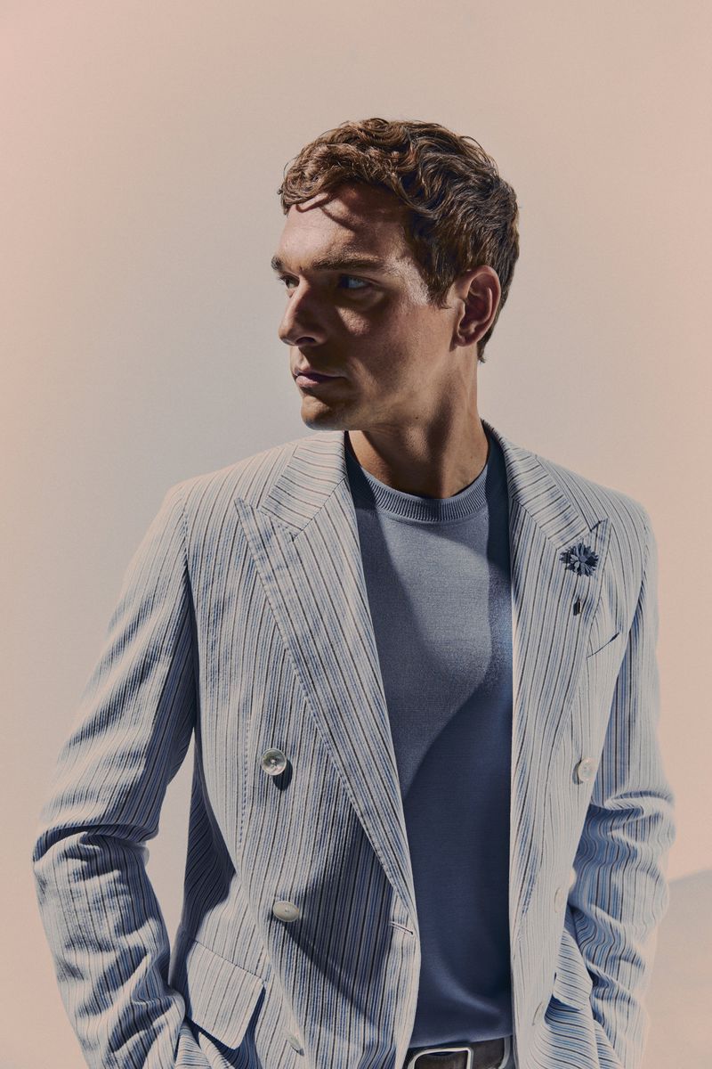 Man in pinstripe blazer and blue t-shirt looking left against a light peach background in studio lighting with shallow depth of field.