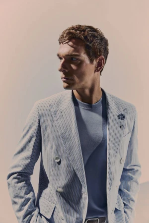 Man in pinstripe blazer and blue t-shirt looking left against a light peach background in studio lighting with shallow depth of field.