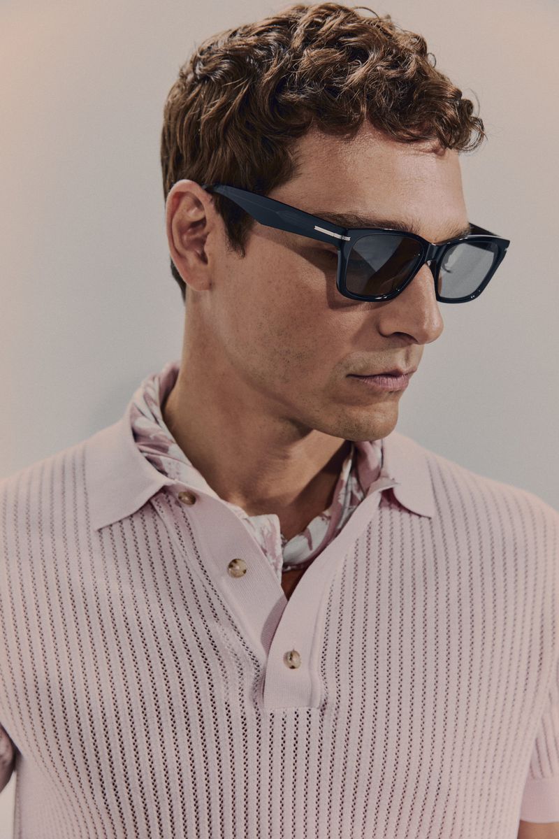 Portrait of a light-skinned adult male wearing dark sunglasses and a pink polo shirt, posing in a photo studio against a neutral backdrop.