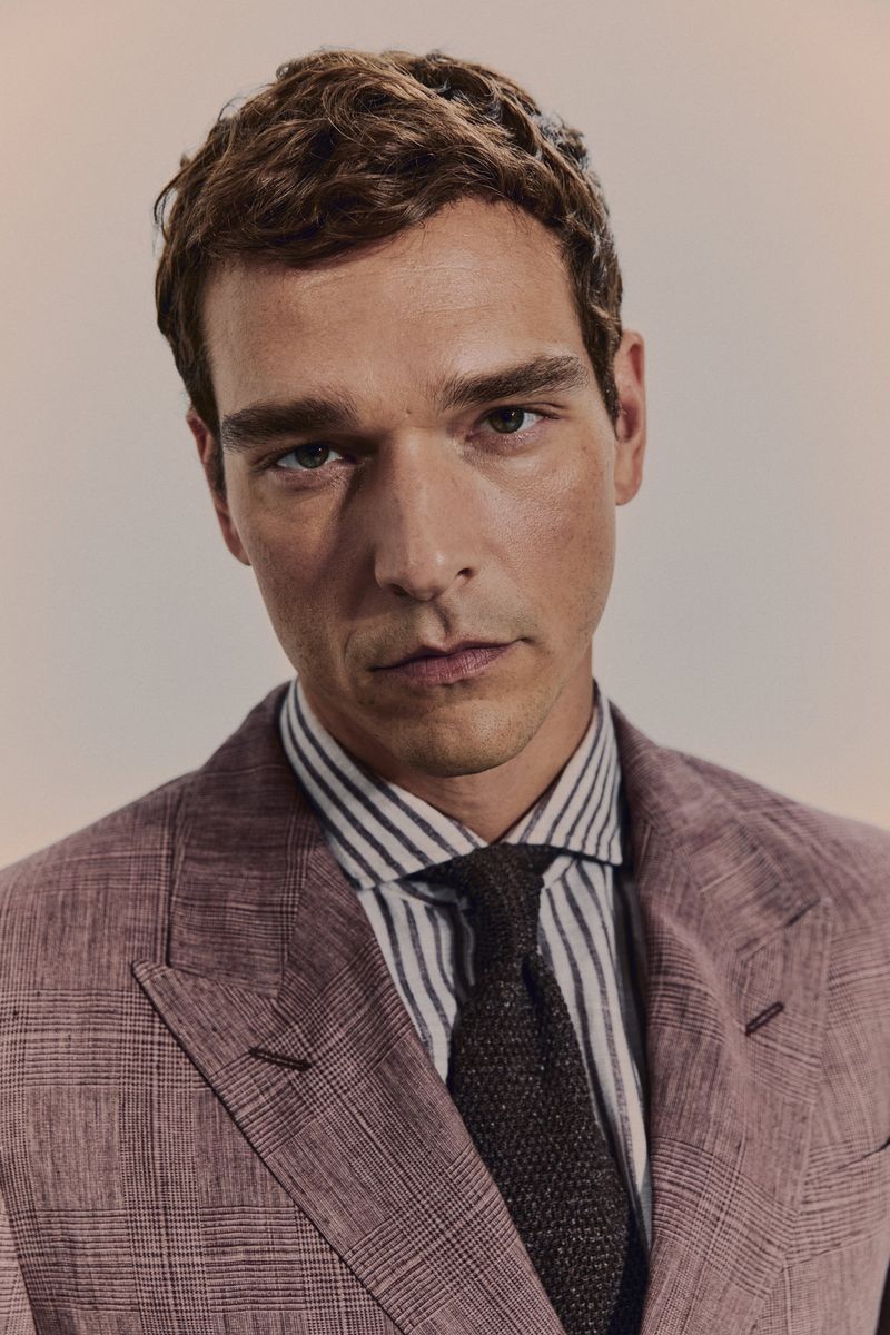 A man in a mauve suit with brown hair gazes forward, his striped shirt and tie complementing the formal yet approachable style.